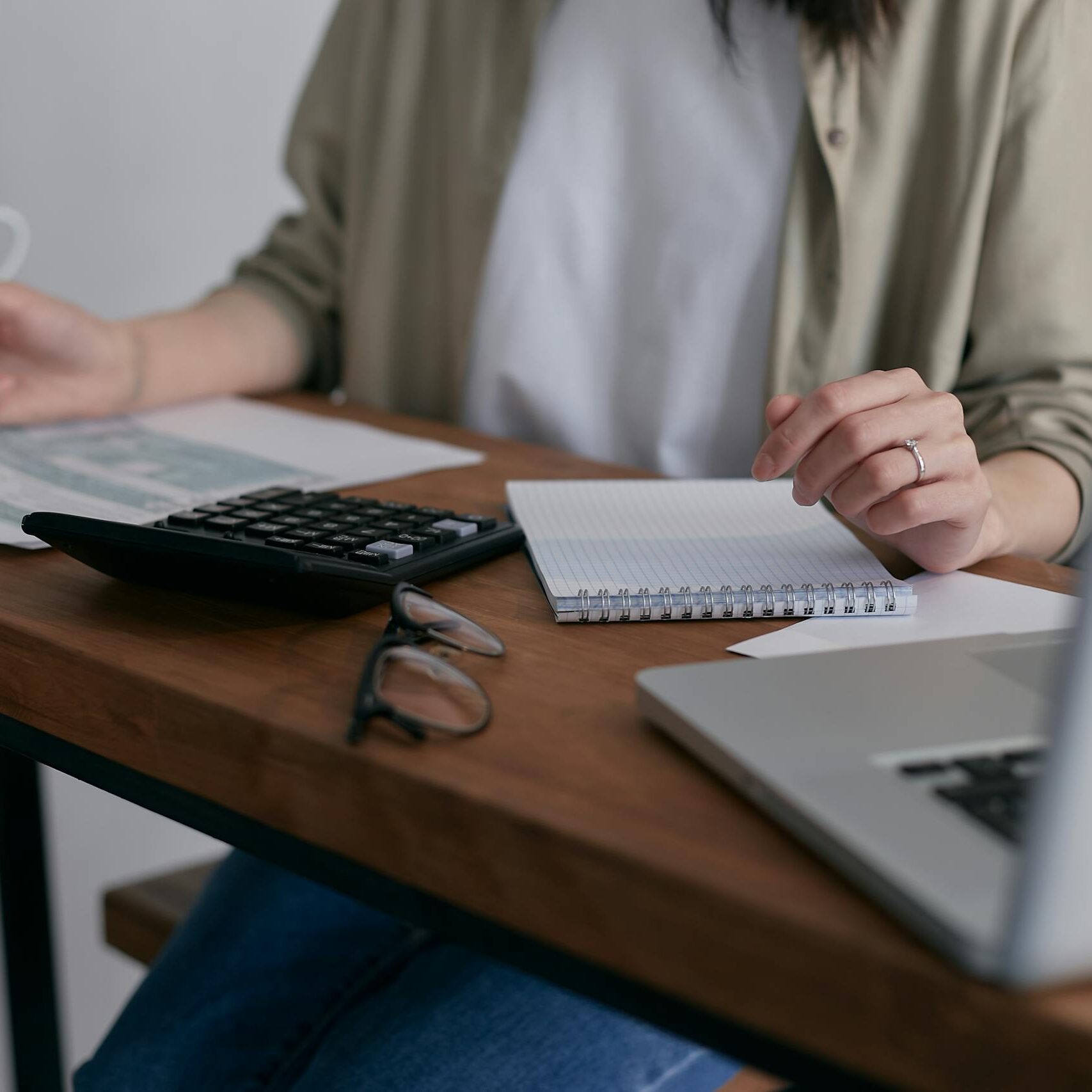 A woman manages finances at home, using a laptop and calculator on a wooden desk.