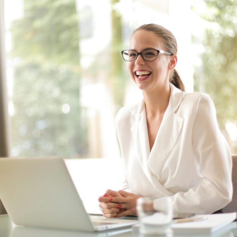 Cheerful businesswoman in glasses working on a laptop, in a bright and modern office setting.