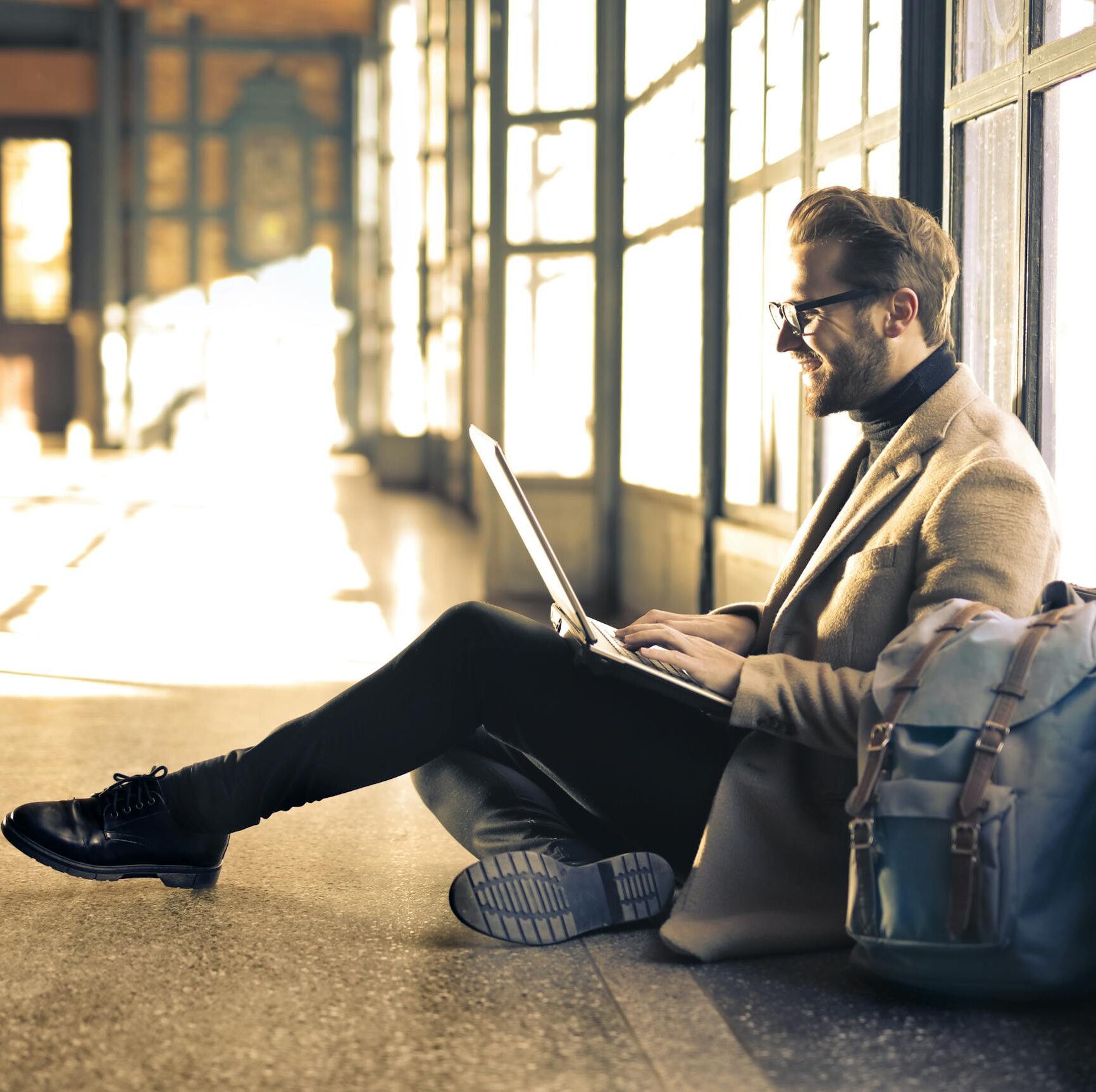 A smiling man sitting by a window in an airport, working on his laptop with sunlight streaming through.